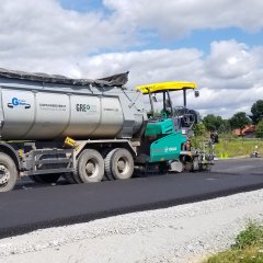 Ein Sattelschlepper steht Fahrbereit vor einem Asphaltfertiger. Im Vordergrund ist frisch hergestellter Asphalt.