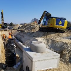 Eine geböschte Baugrube mit einer Tiefe von ca. 2 Meter in der einige Arbeiter zwei Leitungen verlegen. Im Vordergrund sind zwei Schachtbauwerke errichtet worden.