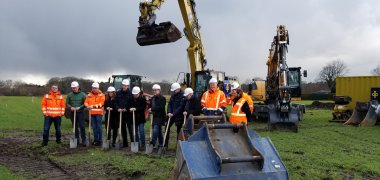 Aufstellung von Menschen und Maschinen für den ersten Spatenstich. Die Vertreter von verschiedenen Projektbeteiligten tragen Helme und haben Spaten in der Hand. 