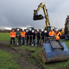 Aufstellung von Menschen und Maschinen für den ersten Spatenstich. Die Vertreter von verschiedenen Projektbeteiligten tragen Helme und haben Spaten in der Hand. 