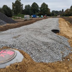 Man erkennt eine Baustelle: Eine zukünftige Straße -  der Schotter wurde gerade eingebaut, jedoch noch nicht verdichtet. Im Vordergrund ist ein provisorisch abgedeckter Schacht.